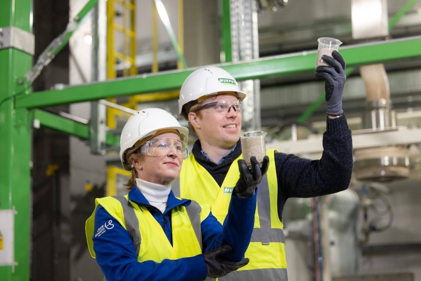 A man and woman, wearing an Uisce Éireann hi-vis jacket holding up a glass of Wastewater sludge at a treatement plant