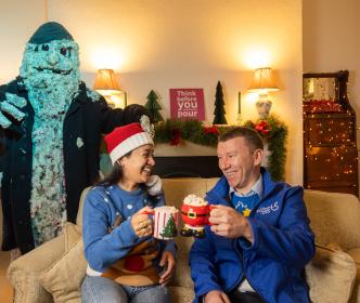 Two Uisce Éireann workers with mugs of hot chocolate and Christmas jumpers on a couch with a scary mascot behind them