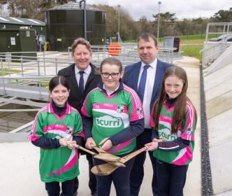 Three girls with jerseys and hurls with two Uisce Éireann workers in a water treatment plant